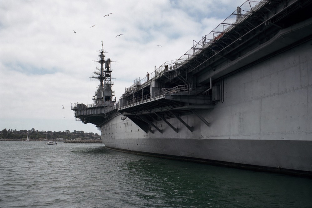 Grey flight deck edge and island tower of the big aircraft carrier in San Diego seen from water level with gulls overhead