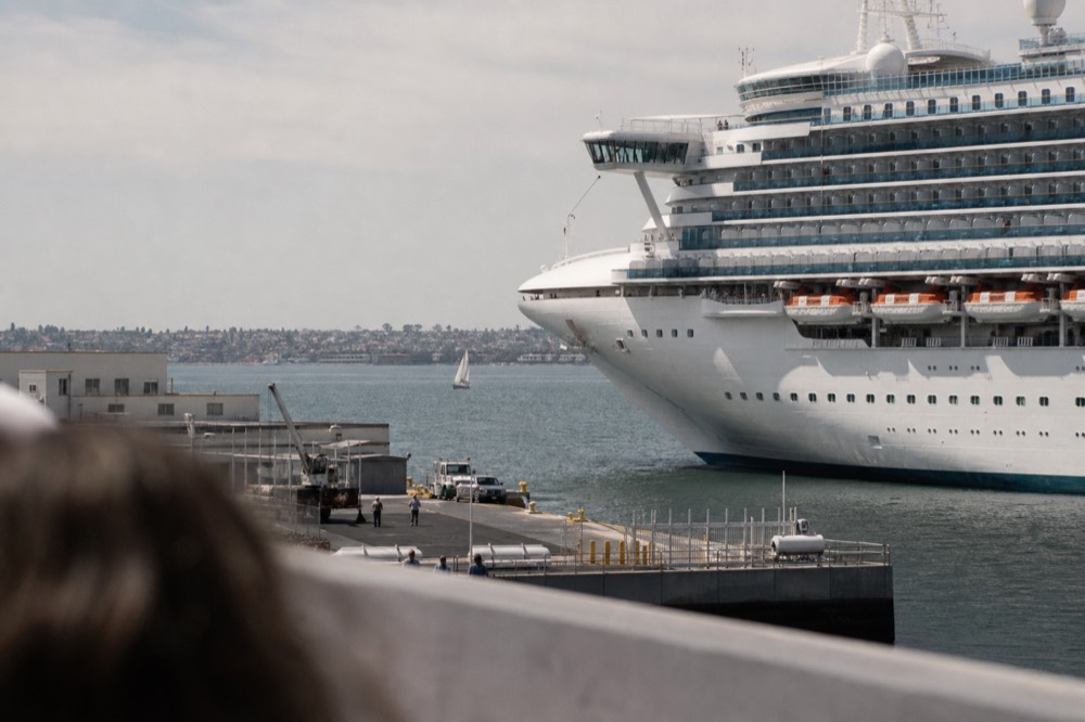 A large white cruise ship at the San Diego Embarcadero terminal, a railing or shoulder softly out of focus in the foreground