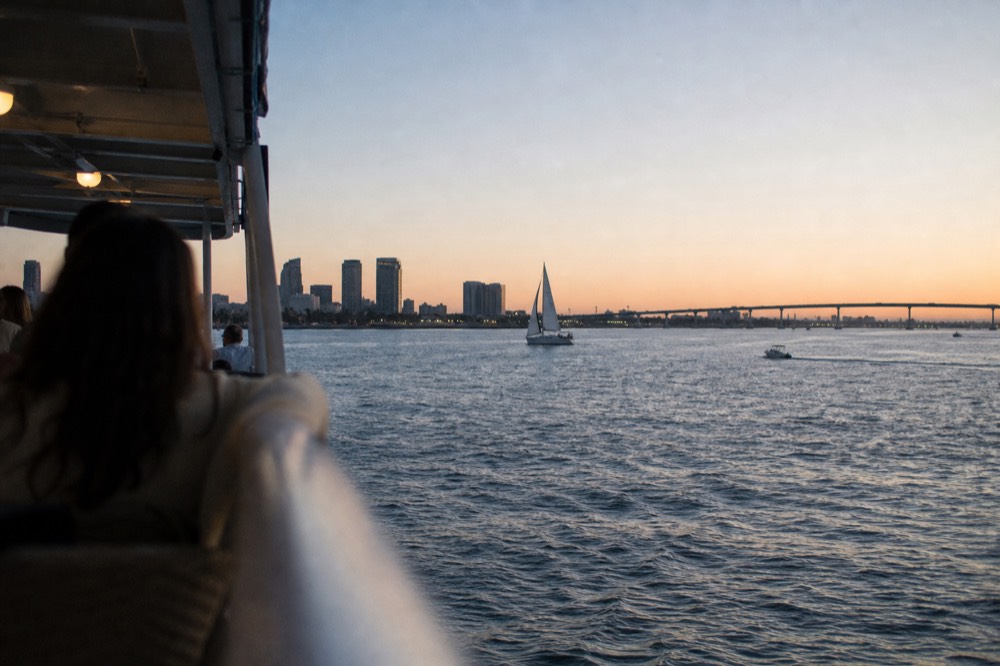 An evening dinner cruise ship on San Diego Bay, a railing or shoulder softly out of focus in the foreground