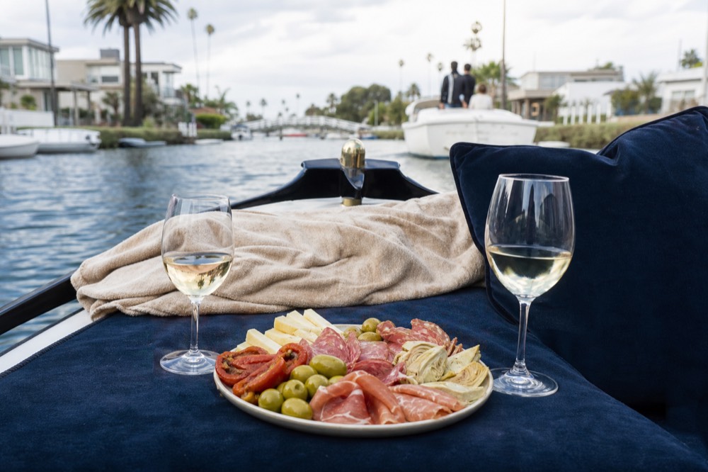 An antipasti plate and two glasses set out on the small table of a Coronado Cays gondola, the still canal water behind