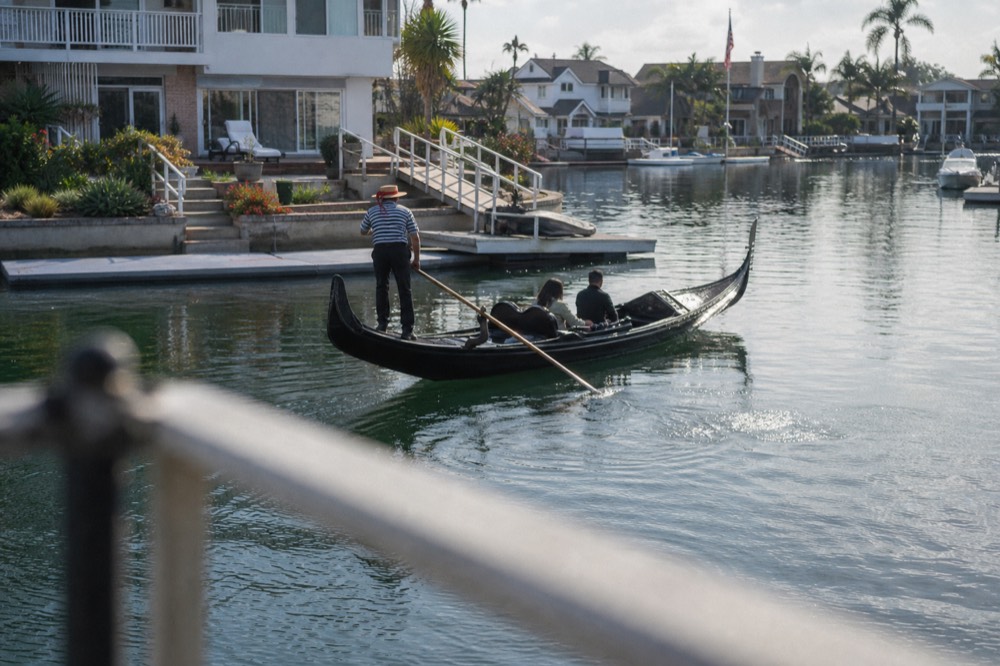A black Venetian gondola in the calm canals of the Coronado Cays, a railing or shoulder softly out of focus in the foreground